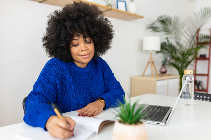 Young latin american student woman taking notes while using laptop at home