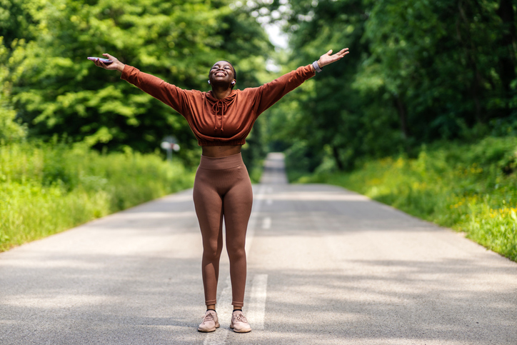 Young active woman in sportswear with raised up hands enjoying beautiful sunshine while taking break after workout outside.