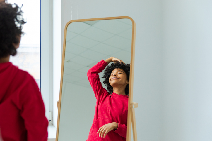 Love yourself. Beautiful young smiling african american woman dancing touching curly hair enjoying her mirror reflection. Black lady looking at mirror looking confident and happy. Self love concept.