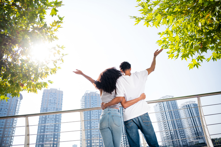 Rear view photo of happy young married people with raised hands walking in modern town looking on sunny sky