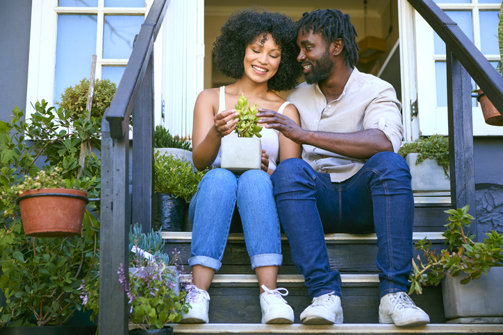 Couple Sitting On Steps Outside At Home Looking After Garden Plants - Blue Zone reference