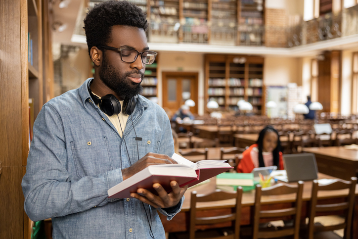 Dark-skinned young man in eyeglasses with a book in hands in the library