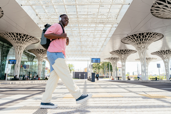 Essential Self-Care Items For Travel - A man wearing a pink shirt and jeans walks down a sidewalk with a backpack. He is listening to music on his headphones