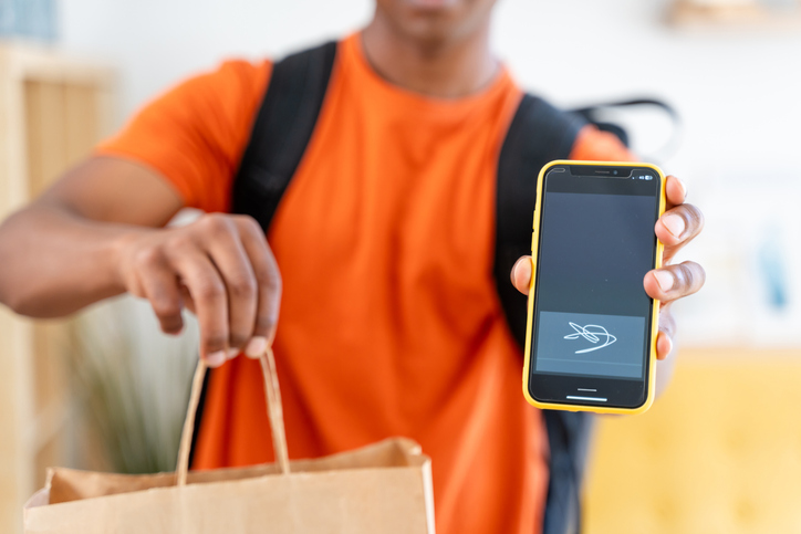 Courier in an orange shirt displays a completed delivery status on a mobile app, holding a paper bag