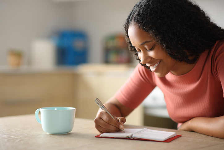 journal spiritual journey - Happy black woman writing in agenda in the kitchen
