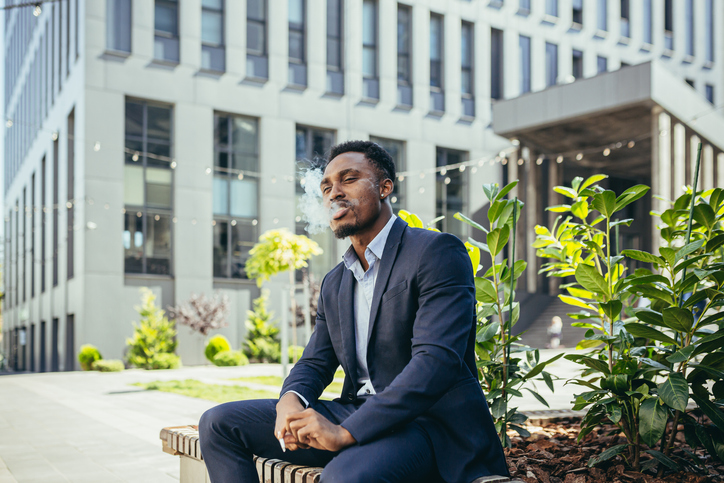 african american business man smoking cannabis outdoors sitting on city park bench on urban street background. Male employee Businessman. Office worker in suit Relieves stress a marijuana outside