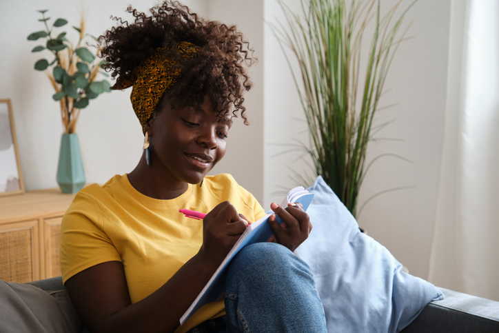 benefits of journaling - Young black woman writing notes sitting on sofa at home