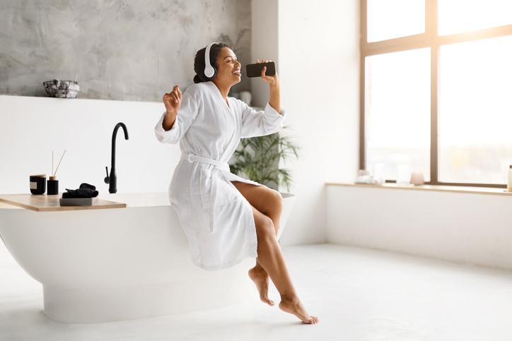 sound bath - Cheerful black woman in bathrobe singing joyfully with smartphone in bathroom