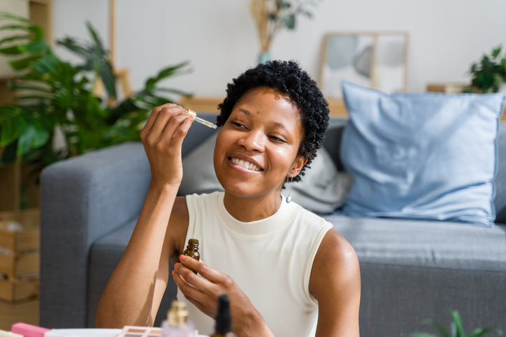 Beautiful black woman applying serum on her face and smiling at home