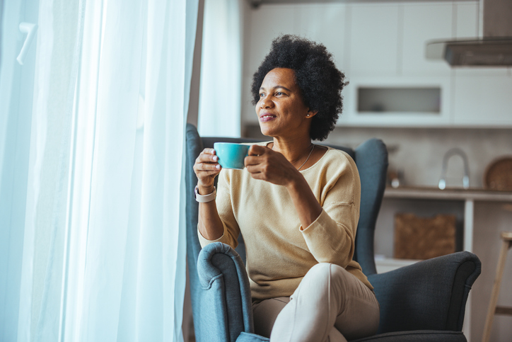 African Woman sitting on her sofa in her living room holding a cup. She relaxing. Portrait of a happy African American woman at home warming up her hands with a cup of coffee