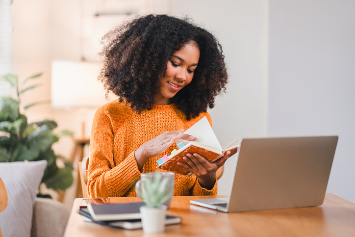 Smiling African American woman reading a notebook while working at home, sitting at a desk with a laptop and a coffee mug.
