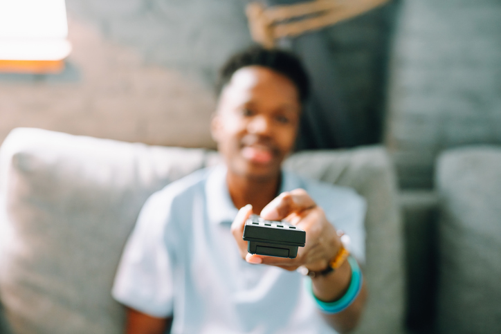 Side Effects Of Watching Too Much TV - Hand with remote control changing channels on TV. Blurred dark skin man and focus on hand