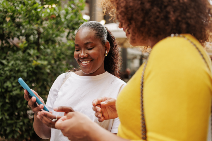 Cheerful african american woman sharing smartphone with friend in city street.