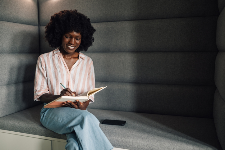 Happy interracial casual businesswoman on sofa writing down in textbook
