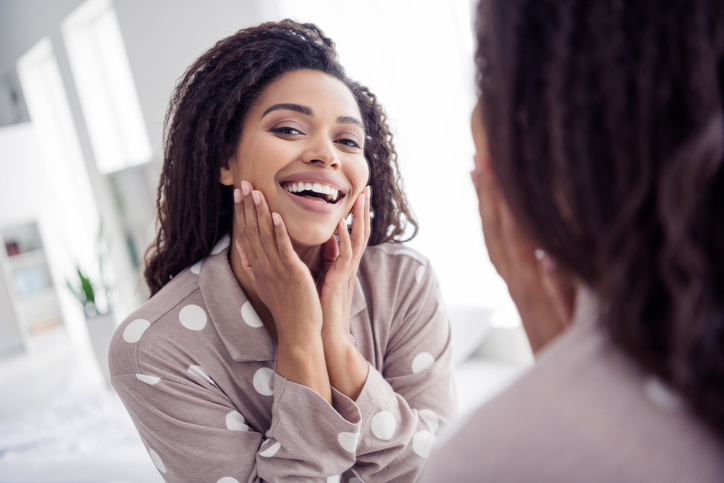 winter skincare - Beautiful woman wearing polka dot pajama smiling joyfully in the mirror at home in the morning, showcasing natural beauty and happiness