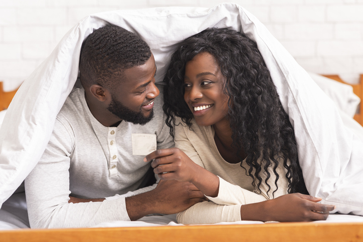 Black couple holding condom, lying on bed under white blanket