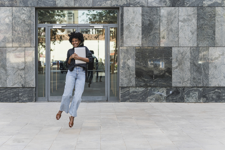 Happy businesswoman holding laptop and jumping in front of office building