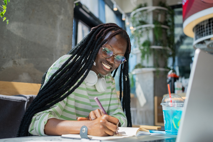 Smiling young African-American woman studying with laptop and notebook in modern cafe.
